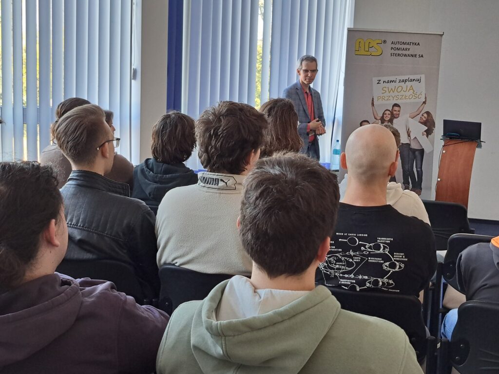 Studenci siedzą w sali konferencyjnej na krzesłach. Pracownik APS opowiada o firmie, zakresie działalności i realizacjach.